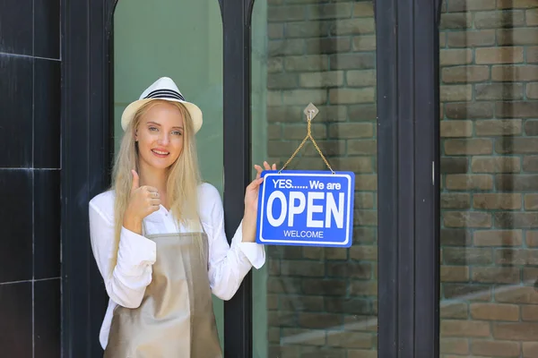 Beautiful blonde hair girl standing by open shop sign plate at door with confidence in front of coffee shop.