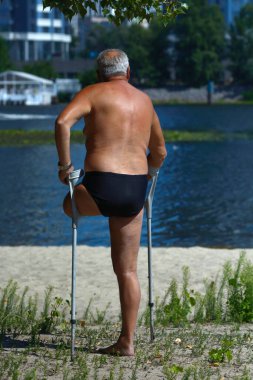 Yaşlı, engelli, tek bacaklı, çift dirsekli koltuk değnekli, kumlu bir sahilde güneşlenen bir adam.