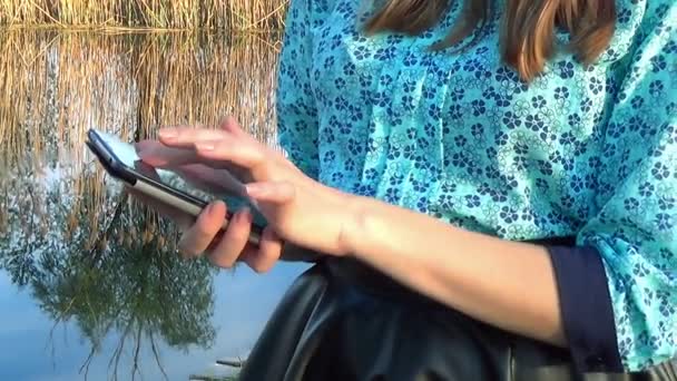 Händerna på en kvinna som använder sin mobil mobiltelefon utomhus av en flod — Stockvideo