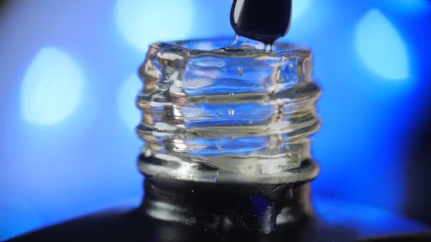 Esmalte de uñas de gel transparente con una botella abierta y una gota goteando en un frasco con un cepillo en el salón de belleza. Fondo azul de la lámpara led. Primer plano. — Vídeo de stock