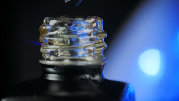 Esmalte de uñas de gel transparente con una botella abierta y una gota goteando en un frasco con un cepillo en el salón de belleza. Fondo azul de la lámpara led. Primer plano. — Vídeos de Stock