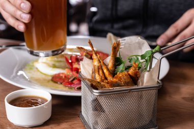 Kızarmış kalamar, domates salatası, kırmızı soğan, kızarmış kırmızı kefal balığı, bir bardak bira ve ahşap masada sos. Bir tabak mürekkep balığının üzerinde çatal ve bira tutan erkeklerin elleri..