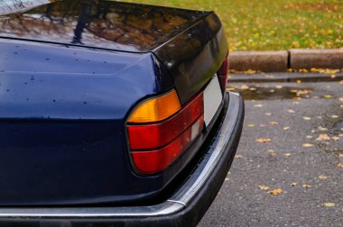 Yağmurlu bir sonbahar parkına park etmiş mavi klasik sedan, sarı yapraklı ıslak ağaçlara karşı kurulmuş ve sakin, nostaljik bir atmosfer yaratan su damlacıklarıyla kaplı.