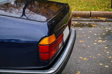 Yağmurlu bir sonbahar parkına park etmiş mavi klasik sedan, sarı yapraklı ıslak ağaçlara karşı kurulmuş ve sakin, nostaljik bir atmosfer yaratan su damlacıklarıyla kaplı.