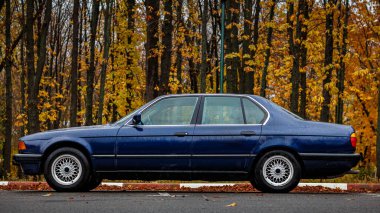 Yağmurlu bir sonbahar parkına park etmiş mavi klasik sedan, sarı yapraklı ıslak ağaçlara karşı kurulmuş ve sakin, nostaljik bir atmosfer yaratan su damlacıklarıyla kaplı.