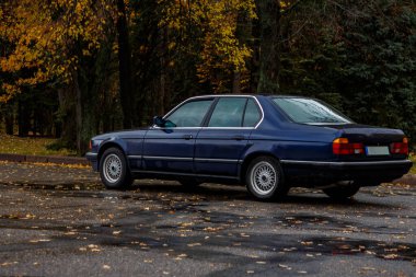 Yağmurlu bir sonbahar parkına park etmiş mavi klasik sedan, sarı yapraklı ıslak ağaçlara karşı kurulmuş ve sakin, nostaljik bir atmosfer yaratan su damlacıklarıyla kaplı.