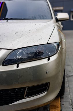 Fenerinde yağmur damlacıkları olan parlak gümüş renkli bir araba, parlak ışıklandırılmış bir oto yıkama istasyonuna yakın plan çekimde yakalandı.