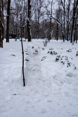 Karla kaplı bir şehir parkında tahta sopa tutan yalnız bir kardan adam: kış aktiviteleri ve çocukluk yaratıcılığı kavramı.