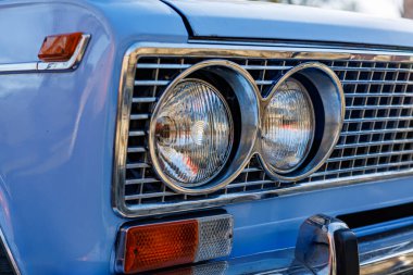 Mavi bir arabanın ön tarafında yakın plan: farlar, krom ızgara ve kaldırım taşlı bir yolda eski beyaz duvar lastikleri..