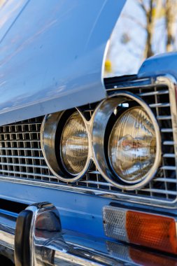 Mavi bir arabanın ön tarafında yakın plan: farlar, krom ızgara ve kaldırım taşlı bir yolda eski beyaz duvar lastikleri..