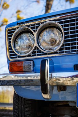 Mavi bir arabanın ön tarafında yakın plan: farlar, krom ızgara ve kaldırım taşlı bir yolda eski beyaz duvar lastikleri..