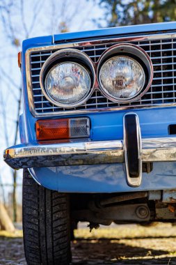 Mavi bir arabanın ön tarafında yakın plan: farlar, krom ızgara ve kaldırım taşlı bir yolda eski beyaz duvar lastikleri..