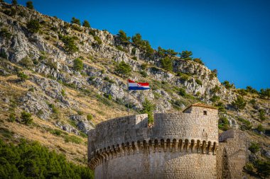 Hırvatistan 'ın Dubrovnik kentindeki bir şato, tepesinde bayrak dalgalanan bir yamaçta bulunuyor. Güneş açık mavi gökyüzünde parlak bir şekilde parlıyor. Ağaçlar dağın alt kısımlarını kaplıyor, manzaranın güzelliğini arttırıyor..