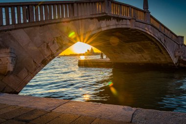 İnsanlar Venedik İtalya şehrinin arkasında güneş batarken köprüde yürür. Gün ışığı söndükçe su gökyüzünün renklerini yansıtır. Sahne, nehrin kıyısındaki hareketli bir alanda sakinliği gösteriyor..