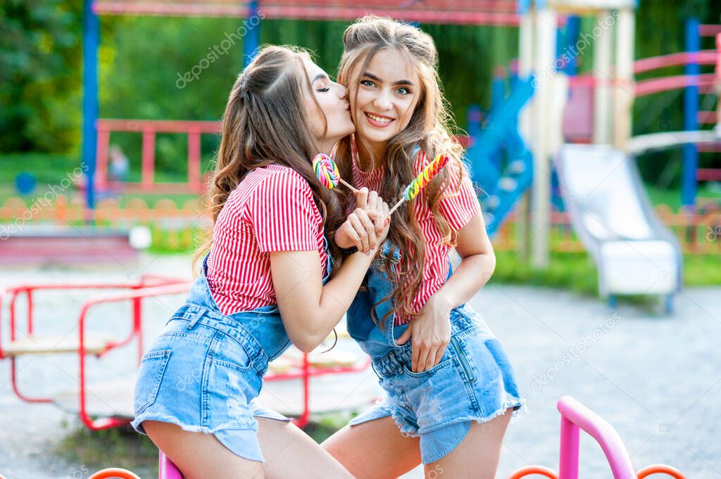 two happy twin sisters laughing and kissing in denim overalls with lollipops on the playground