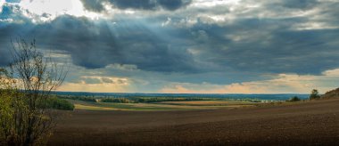 beautiful panorama of plowed land with fields, forests on the horizon, sun rays through the dark sky