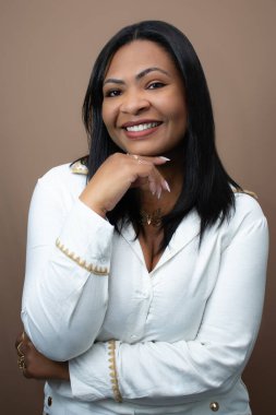 Eli çenesinde, gülümseyen ve kameraya bakan güzel siyah kadın. Kahverengi arkaplanlı stüdyo portresi, güzellik kampanyaları, güven, güçlendirme ve pozitif kadınsı temalar için ideal.