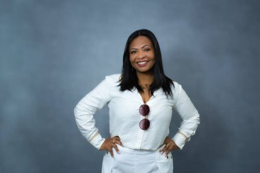 Yakasından sarkan güneş gözlüklü, kameraya gülümseyen beyaz elbiseli çekici siyahi kadın. Fotokopi alanı, kadın modası, kadınlık ve vücut pozitifliği için ideal yatay stüdyo portresi..