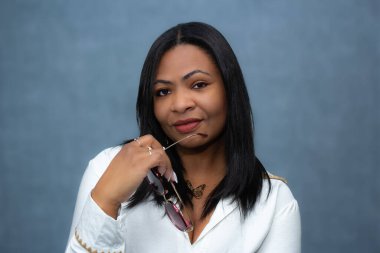 Beyaz elbiseli, elinde güneş gözlüğü olan ve kameraya bakan güzel siyahi bir kadının vesikalığı. Fotokopi alanı olan yatay stüdyo portresi, zengin yaşam tarzı, kadın gücü, kendine güven için ideal.