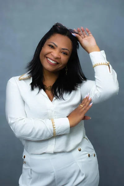 Kafasının üstünde güneş gözlüğü olan beyaz elbiseli neşeli siyah kadın kameraya gülümsüyor. Zengin yaşam tarzı, yaz atmosferi, tatil gezisi ve mutluluk için ideal dikey stüdyo portresi.