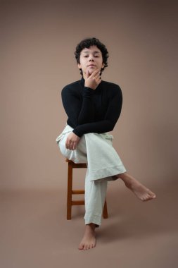 Kısa kıvırcık saçlı güçlü genç bir kadın bacak bacak üstüne atmış ve çenesi üstüne düşmüş bir taburede oturuyor. Fotokopi alanı olan bir stüdyo çekimi, Gen Z için mükemmel bir tutum, kadın gücü ve kadınsılık..