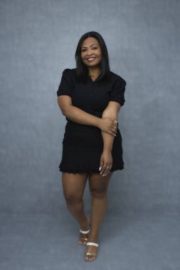 Siyah kıyafetli çekici bir Brezilyalı kadın, zarif bir şekilde poz veriyor ve kameraya gülümsüyor. Kopya alanı ile tam boy stüdyo portresi, özgüven, güven ve kadın güçlendirme kavramları için ideal..