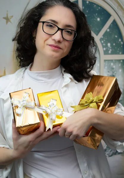 Gözlüklü, beyaz elbiseli, gülümseyen ve elinde üç tane altın Noel hediyesi olan bir kadın. Stüdyo portresi Noel hediyeleri, bayram kutlamaları ve kış mevsimi reklamları için mükemmel..