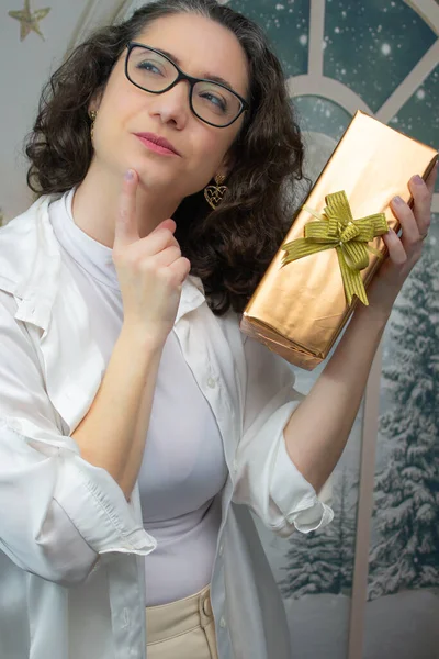 Esmer kadın elinde bir hediye tutuyor ve tatil sezonunu düşünüyor. Yılbaşı hediyeleri, Noel hediyesi alışverişi ve kış mevsimi hayal teması için ideal belden yukarı stüdyo portresi.