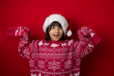 Smiling girl wearing a Santa hat and Christmas sweater raising her hands happily on a red background with copy space, perfect for festive designs and holiday campaigns