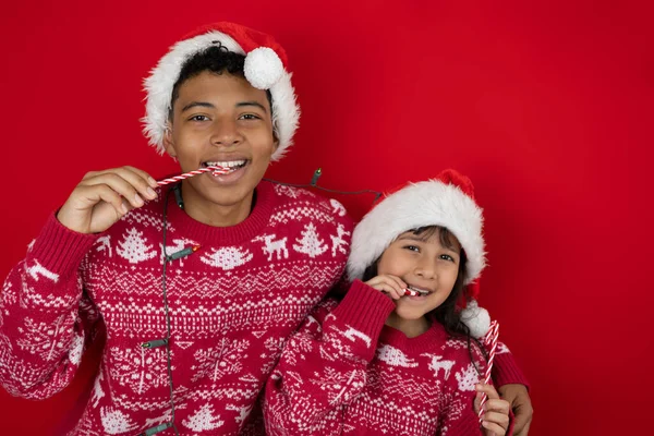 Brown skinned girl and teenager wearing Christmas hats and sweaters enjoy eating candy canes and peppermint candies on a red background. Cheerful and festive image ideal for promotions, ads, or Christmas designs