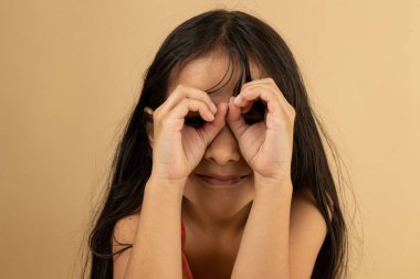 Elleriyle dürbün yapan ve şefkatle bakan gülümseyen genç bir kıza yaklaş. Fotokopi alanı olan açık kahverengi arkaplan, reklam veya tasarım için ideal