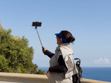 Güneş gözlüklü esmer kadın, güneşli bir günde arka planda mavi gökyüzü ve deniz olan selfie çubuğuyla telefonuyla fotoğraf çekiyor. Turizm ve yaz için ideal bir görüntü.