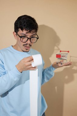 Elinde gagası ve minyatür kırmızı alışveriş arabası olan bir adam, şaşırmış görünüyor, ev giderleri kavramı ve artan yaşam maliyeti