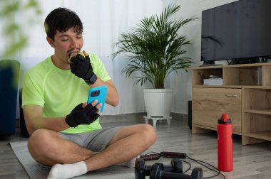 Oturma odasındaki paspasın üzerinde oturan genç bir sporcu telefonuna bakarken elma yiyor. Sağlıklı yaşam tarzı, teknoloji ve ev egzersizi kavramı