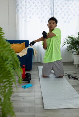 Latin genç bir adam paspasın üzerinde diz çöküp evinde ısınmak için elini esnetiyor. Hareket kabiliyetini geliştiriyor ve vücudunu egzersize hazırlıyor.