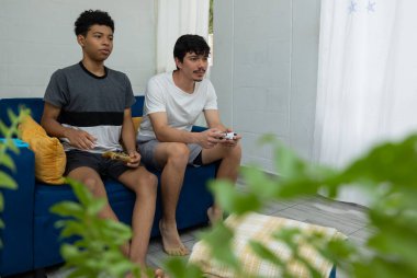 İki arkadaş kanepede oturmuş video oyunu oynuyorlar. Ev eğlencesi sahnesi, arkadaşlar arasındaki dijital rahatlığı, sosyal etkileşimi ve eğlenceyi yansıtıyor.