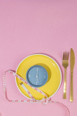 Sarı plaka, saat ve ölçüm bandıyla üzerine yerleştirilmiş, fotokopi alanı, aralıklı oruç kavramı, ağırlık kontrolü ve sağlıklı beslenme ile birlikte pastel pembe arka planda bulunan çatal bıçak takımı.