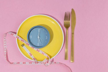 Sarı plaka, saat ve ölçüm bandıyla üzerine yerleştirilmiş, fotokopi alanı, aralıklı oruç kavramı, ağırlık kontrolü ve sağlıklı beslenme ile birlikte pastel pembe arka planda bulunan çatal bıçak takımı.