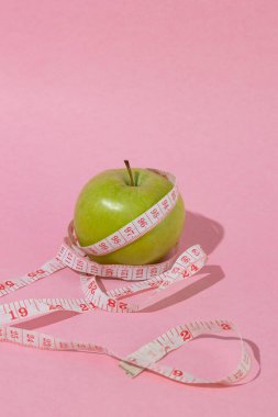Fotokopi alanı, kilo kontrol konsepti, sağlıklı beslenme, dengeli beslenme ve sağlıklı yaşam tarzı olan pastel pembe arka planda ölçüm bandıyla sarılı yeşil elma.