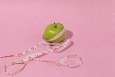 Fotokopi alanı, kilo kontrol konsepti, sağlıklı beslenme, dengeli beslenme ve sağlıklı yaşam tarzı olan pastel pembe arka planda ölçüm bandıyla sarılı yeşil elma.