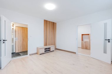 Geniş, yenilenmiş, ahşap kaplamalı zemin, beyaz duvarlar, ahşap kaplamalı aksan duvarı ve alçak medya dolabı, iki kapı, döşemeye hazır parlak minimalist alan.