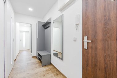 Daire girişi koridorunda ahşap bir zemin, beyaz duvarlar ve dolabı da dahil olmak üzere minimalist gri bir mobilya seti, ayakkabı askısı, ceket askısı ve ayna var.