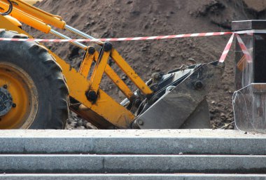 Elinde kova olan traktör, kırmızı ve beyaz renkli koruyucu bandın arkasında çalışıyor. Yol yapım ve onarım çalışmaları