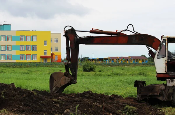 An excavator digs a ditch in a vacant lot against the backdrop of a colorful house. Pipeline laying. reportage shooting. — Stock Photo, Image