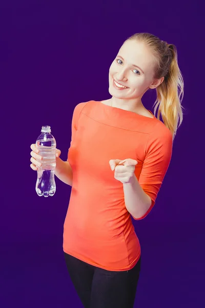 Donna sorridente che mostra l'indice alla fotocamera mentre tiene in mano una bottiglia d'acqua segno in piedi su uno sfondo blu — Foto Stock