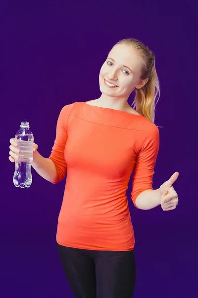 Donna sorridente che mostra un segno pollice in su mentre tiene in mano una bottiglia d'acqua mentre in piedi su uno sfondo blu — Foto Stock