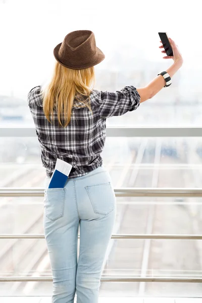 Młoda kobieta odwróciła się plecami i stoi na tle szklanej ściany i robi selfie. kieszeń paszportu i biletu — Zdjęcie stockowe