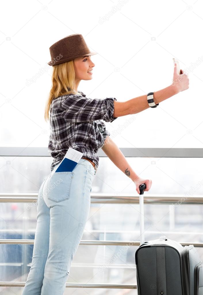 woman in a hat and a suitcase standing on the background of glass wall and makes selfie. pocket a passport and a ticket.