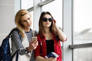 Terminal bir bavul, bir sırt çantası ve bir pasaport ile ayakta iki güzel kadın telefona bakmak