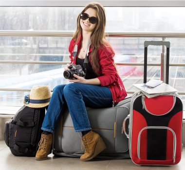 genç çekici kadın kırmızı bir ceket ve bavul terminal veya tren istasyonunda oturan güneş gözlüğü.